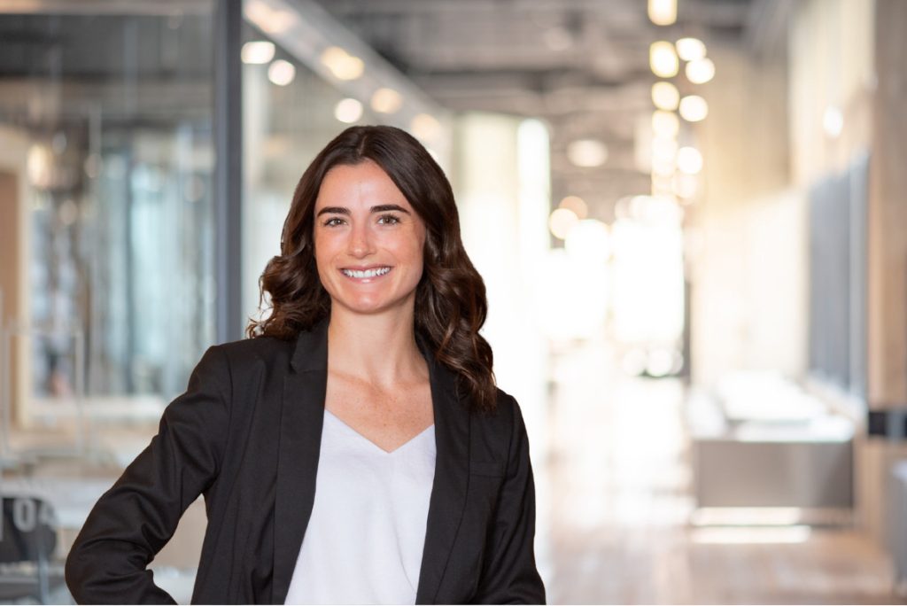 A woman with long brown hair in a black blazer and white shirt stands smiling in a modern office hallway.