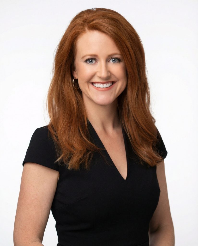 Woman with long red hair, wearing a short-sleeved black dress, smiling at the camera against a plain white background.